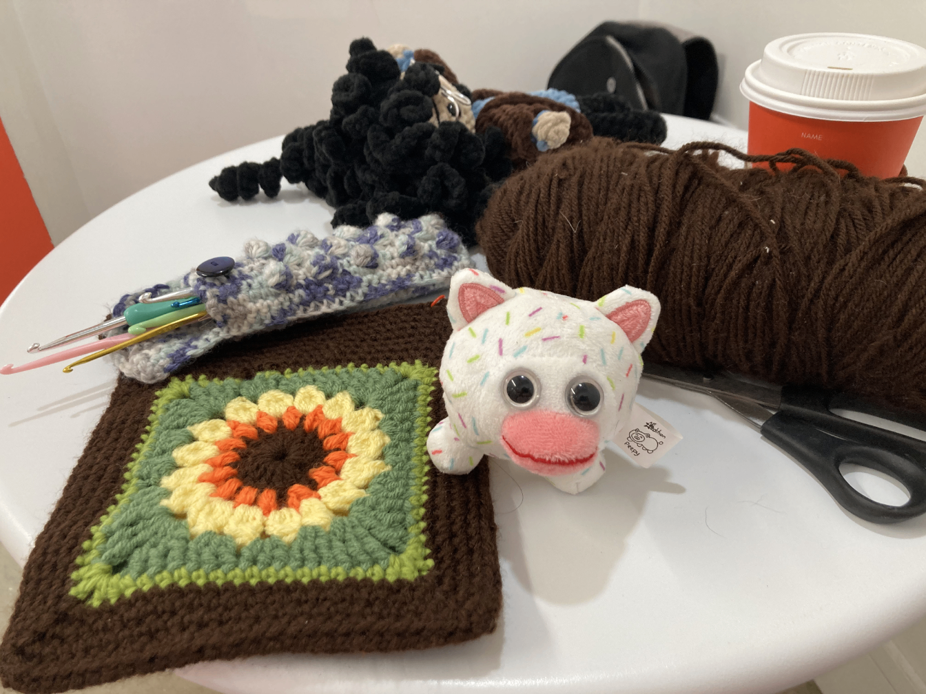 An indoor coffee shop table with a bunch of stuff on it. Contents include a large ball of brown yarn, scissors, a coffee cup, my crocheted sunflower bag, my open crocheted hook case, my friend's crocheted doll and my Icecreampy plush.
