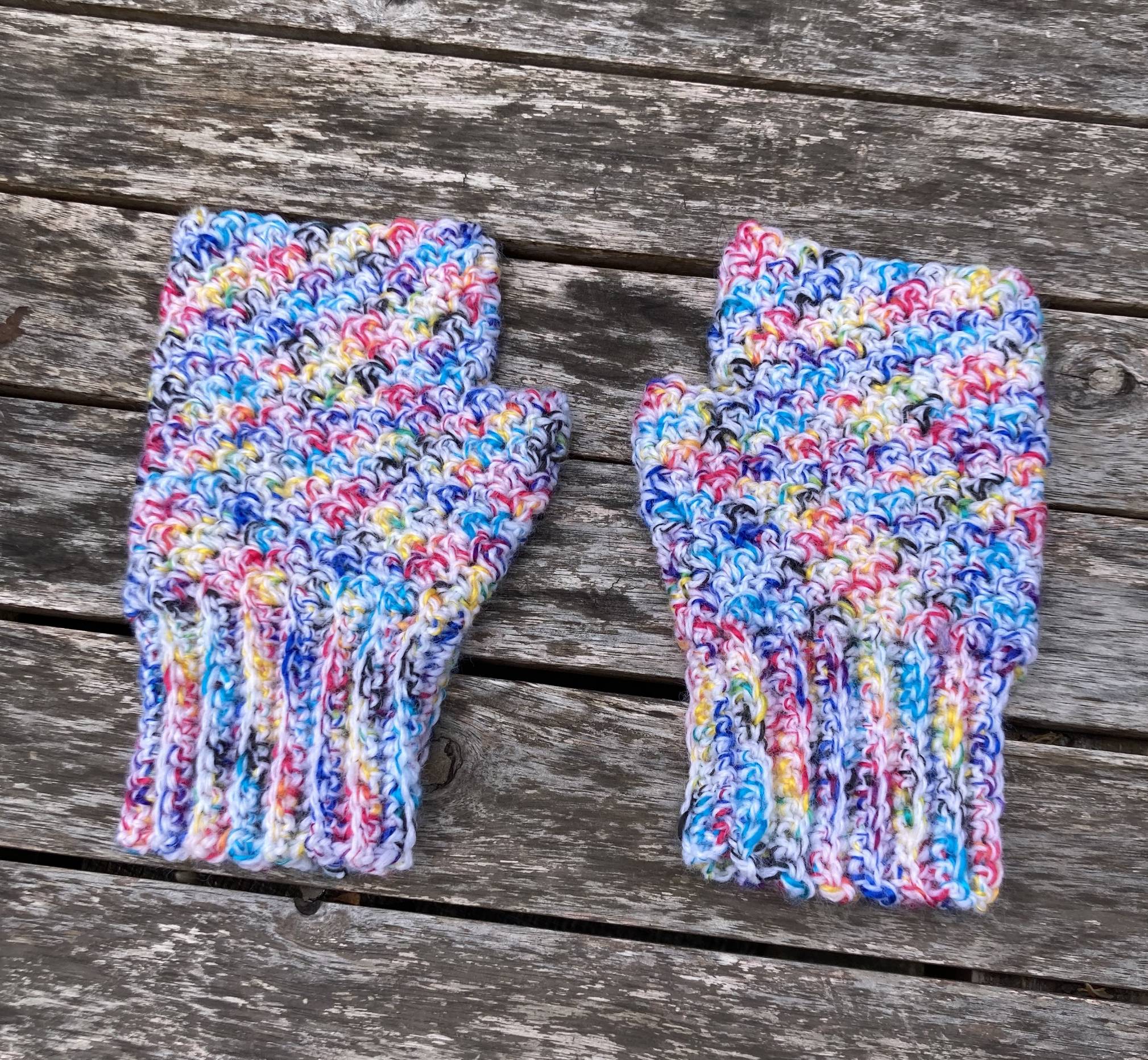 A pair of crocheted fingerless gloves made of white and rainbow yarn on a table.