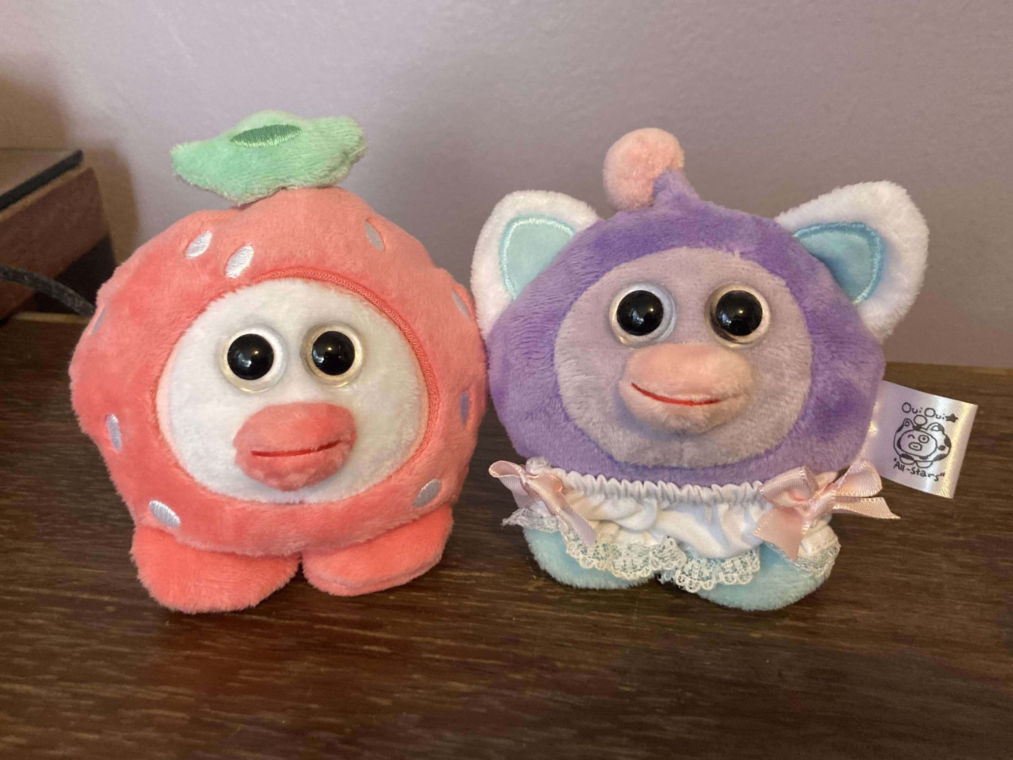 A OuiOui wearing a strawberry shaped outfit that covers its whole body and a purple OuiOui wearing frilly underpants side by side.